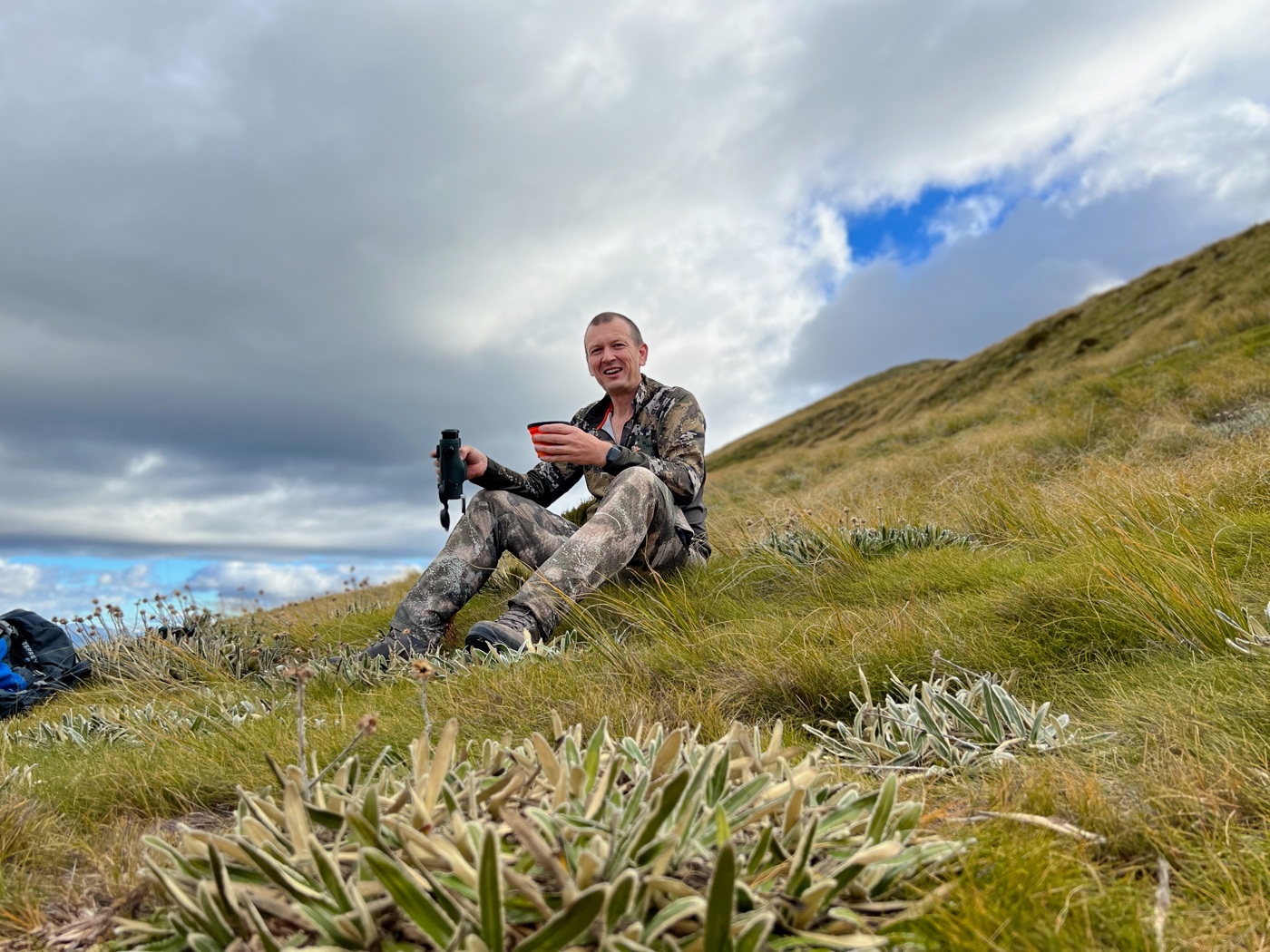 the developer sitting in tussock on an open face with a rangefinder in one hand and a brew in the other, under a heavy sky.