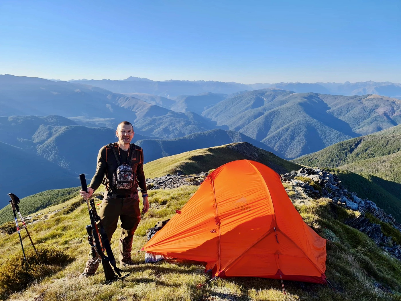 The developer beside a small orange tent camped on a ridge top, rifle in hand, with the Southern Alps behind him.